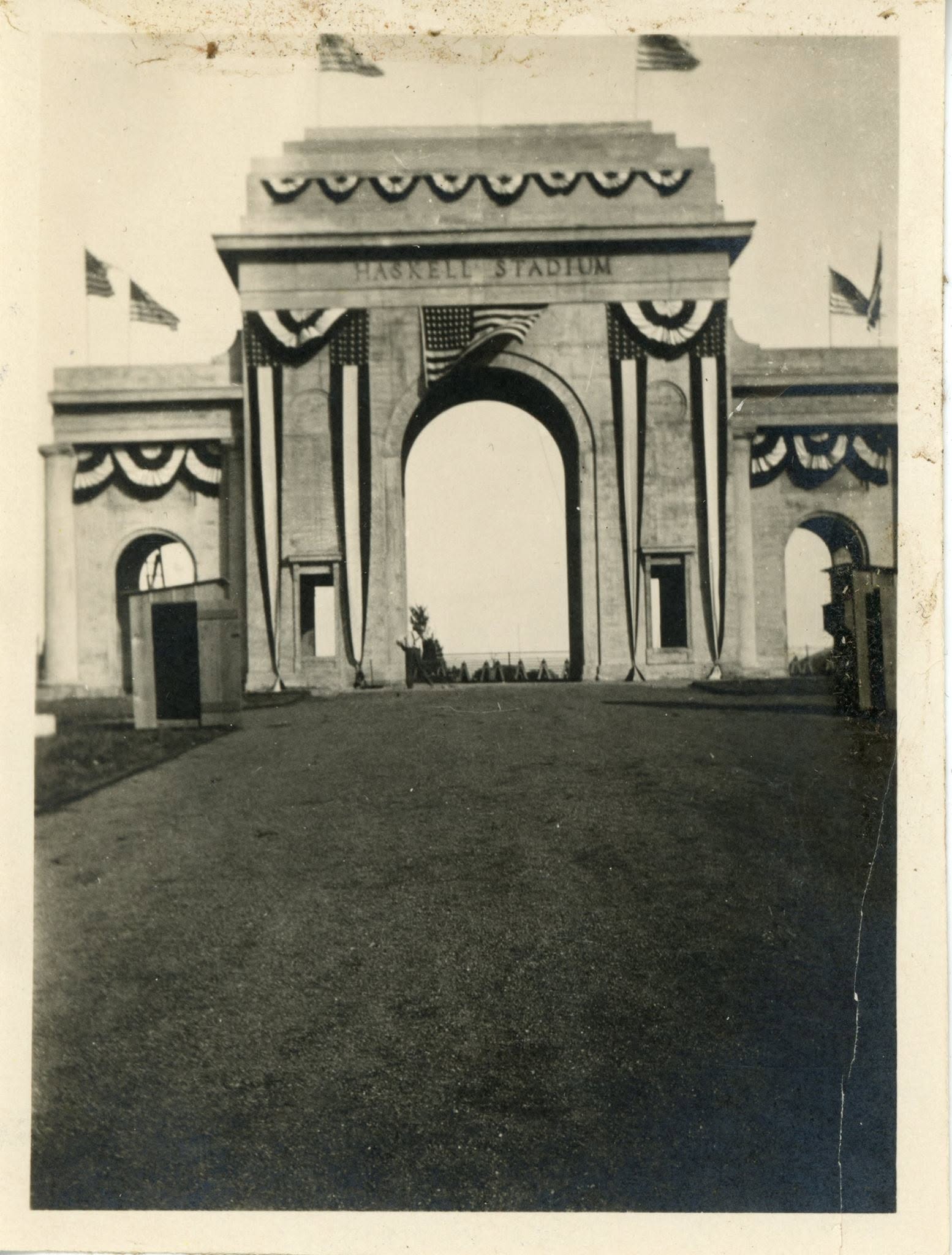 A icolorless mage with grass covering the foreground of the image. In the background, an arch stands with two smaller arches connected on each side. A total of six American flags wave, with two on each of the arches. Banners line the top of each arch. On the middle arch are the words "HASKELL STADIUM"